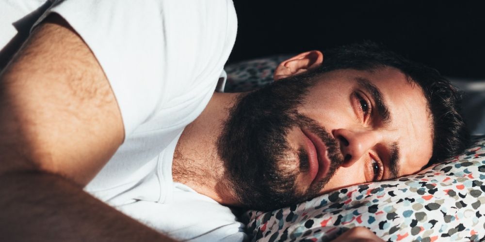 Erschöpfter Mann liegt mit Schlafproblemen im Bett – Symbolbild für den Online-Selbsttest bei Schlafproblemen