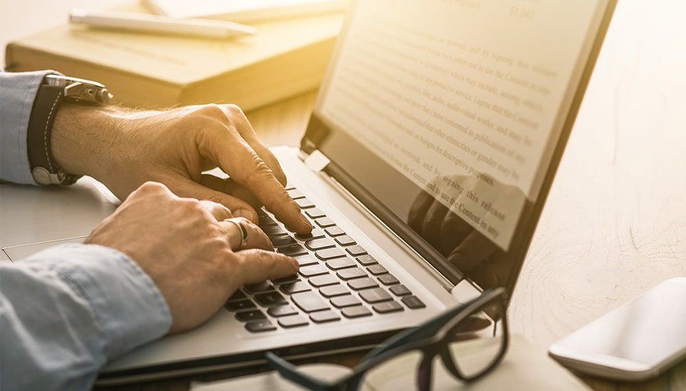 Person arbeitet an einem Laptop, daneben liegen Brille, Smartphone und Bücher – symbolisiert digitales Recherchieren oder Schreiben im Kontext von mentaler Gesundheit.
